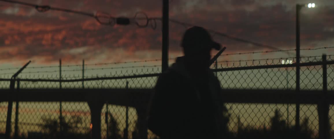 The silhouette of a man walking past a chain link fence with barbed wire ontop