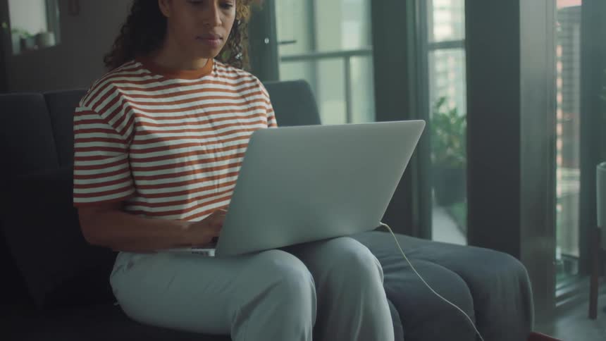 A teenage girl sits on a couch at home and moves to music as she uses her laptop