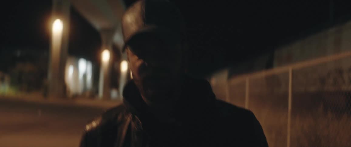 A man wearing a cap is walking next to a fence at night and he looks behind him as a train passes on the other side of the fence