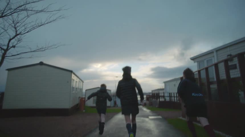 Three boys run through a trailer park while tossing a soccer ball