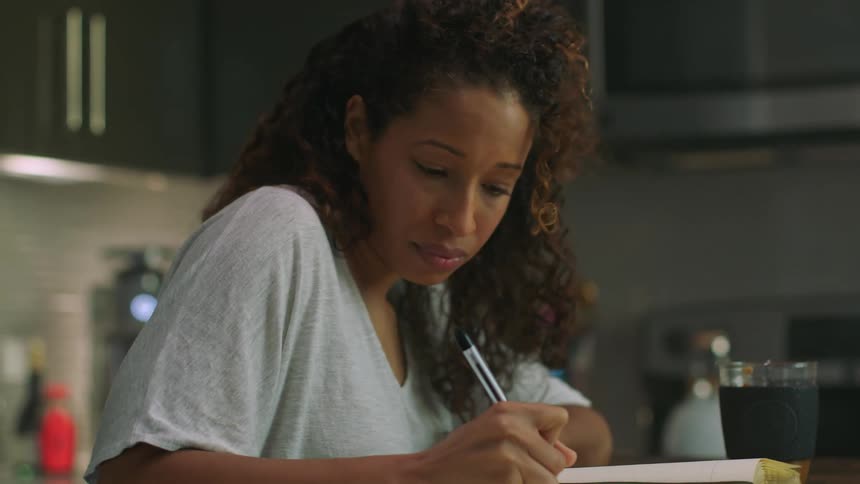 Woman with curly hair and white shirt writes in notebook sitting at table in kitchen