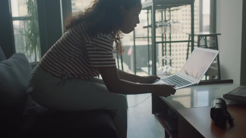 A woman at home walks in to a room picks up her laptop and sits on the couch and puts her feet up