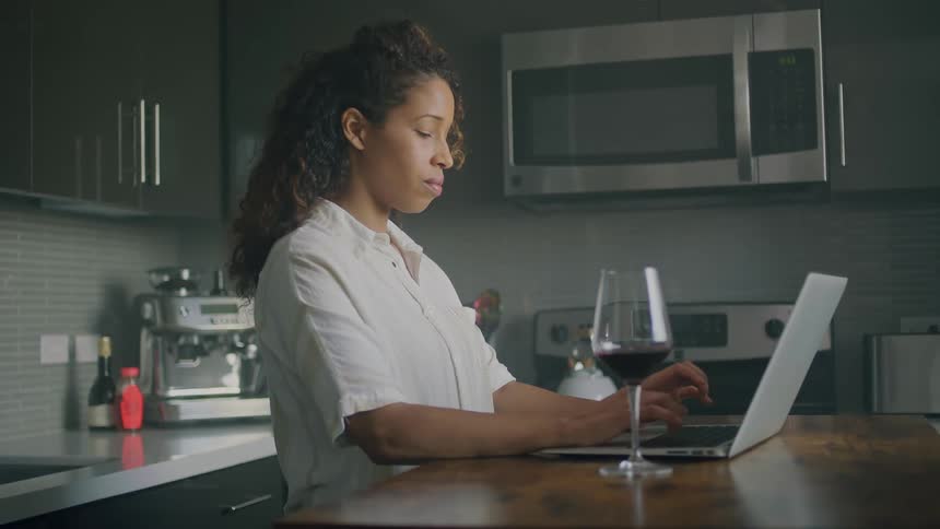 Woman with curly hair and white shirt uses laptop on kitchen island next to a glass of wine