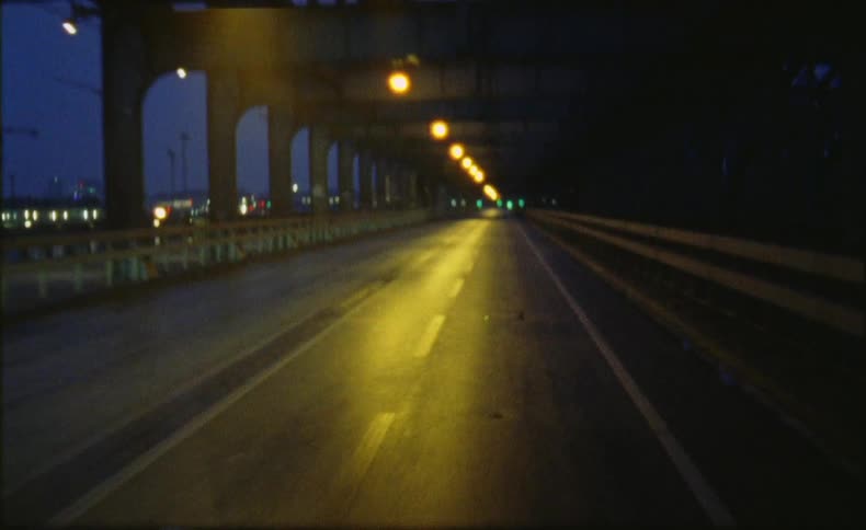 Yellow lights glow on a bridge road with city lights and construction cranes in the distance