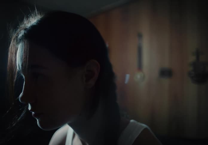 A woman sits with her head bowed in a dim room with wood paneling
