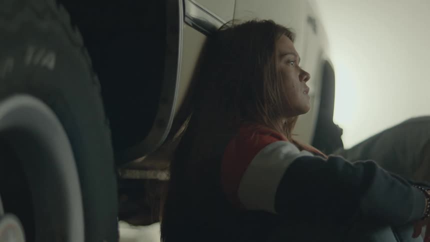 Two young adults sit together on the floor leaning against a vehicle in a depressed state