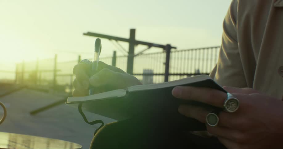 A man is sitting outside on a rooftop with a cup of coffee and writing in a book