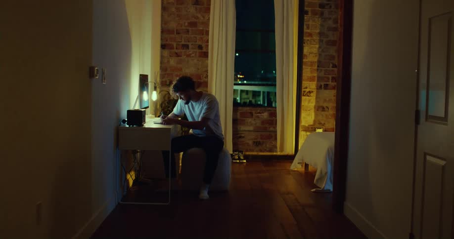 Man sits at table writing as nearby window shows semi truck passing on overpass