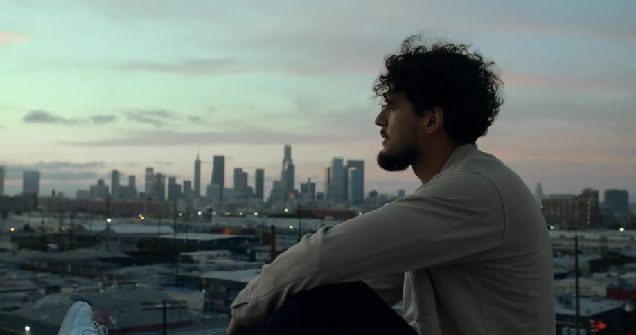 A man looks out over a cityscape while sitting on the edge of a rooftop in the evening