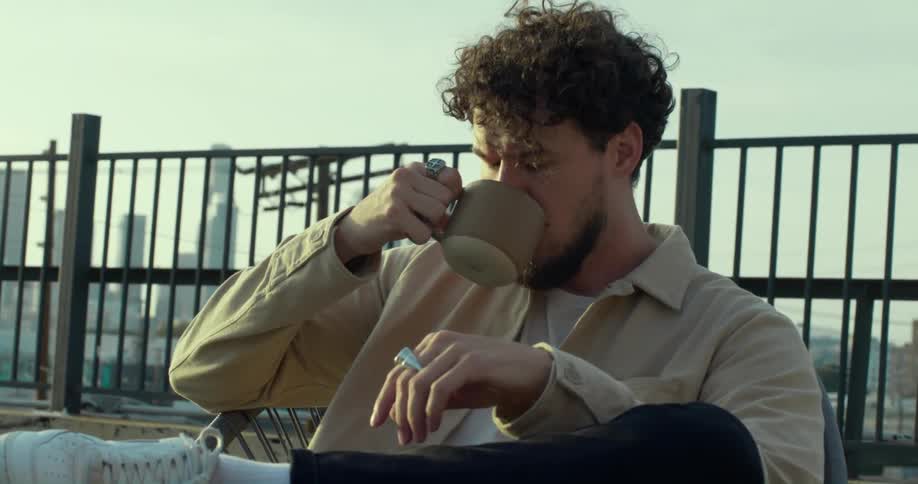 A man sits on a rooftop with the city skyline behind him and drinks coffee
