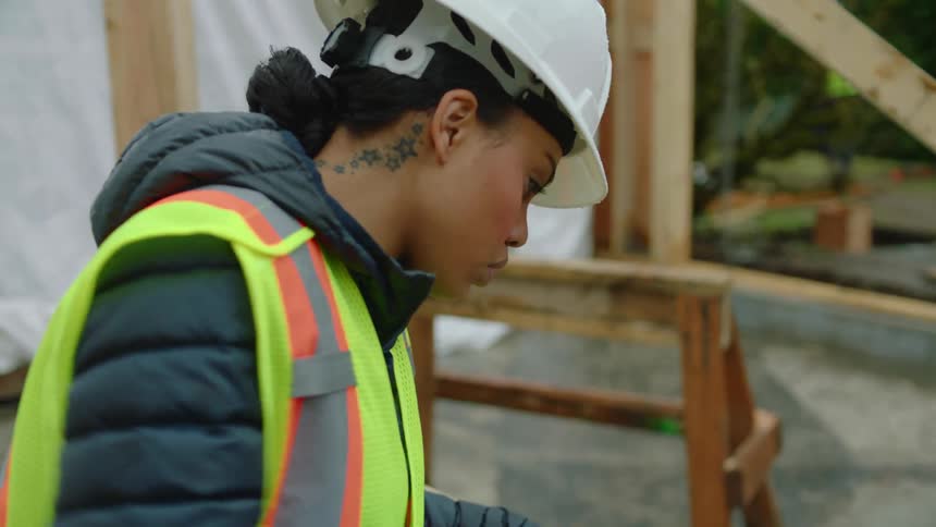 A female construction worker is sawing wood as another crew member walks around the site