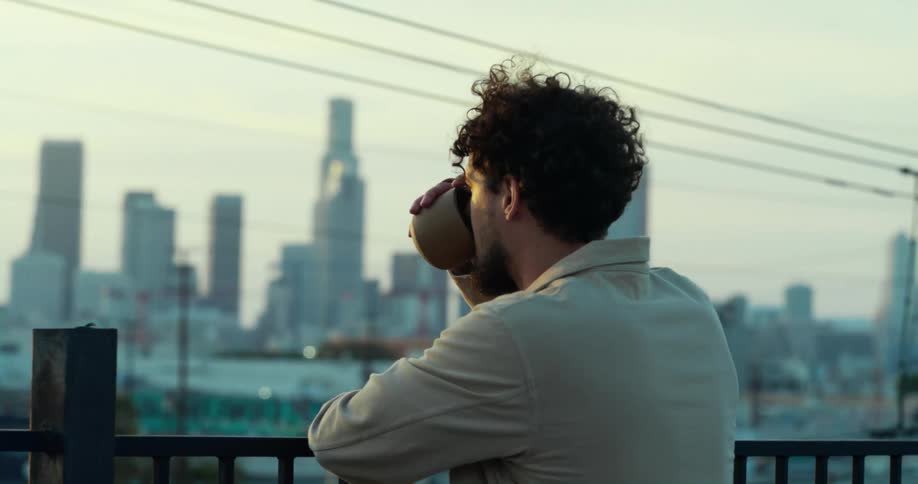 A man leaning against a railing drinking his morning coffee looking out at the city skyline
