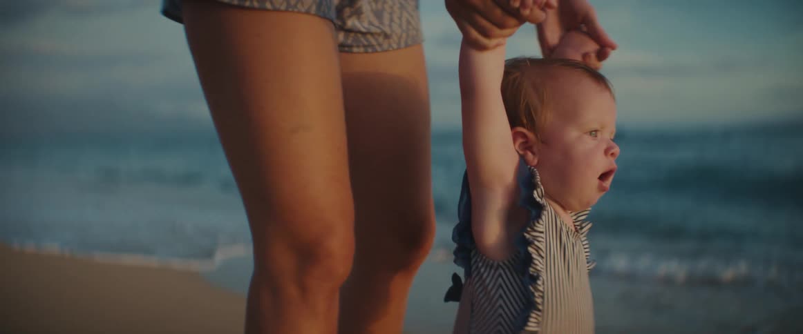 A woman holds her babys arms up so she can walk along the beach