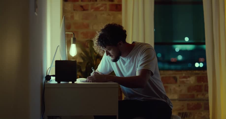 A man sits at a small desk writing by a small lamp