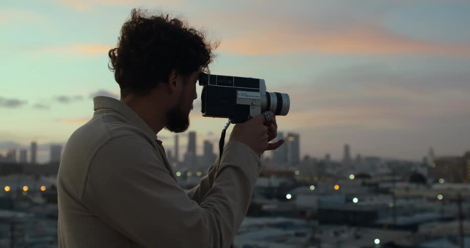 A young man standing on a building on the outskirts of downtown brings a vintage video camera up to his eye and looks through as the wind blows through his hair