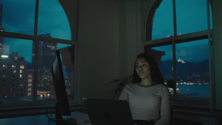 A woman sits at a desk in a dark room frustrated with a computer in front of her