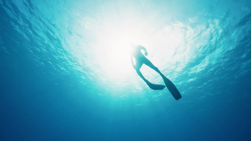 Silhouette of a person scuba diving in clear water as the sun shines above