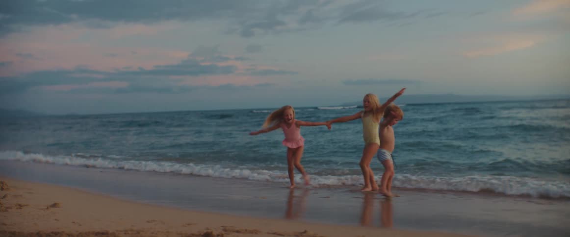 Three young children are playing on a beach as the waves roll in and the little boy falls in the sand