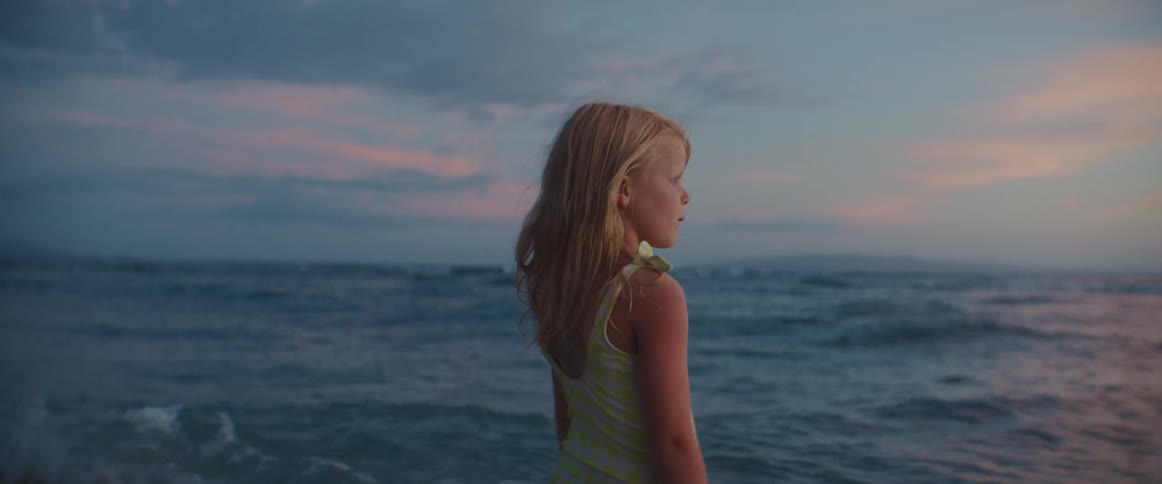 A little girl with long blond hair is standing near the water on a beach as the waves roll in