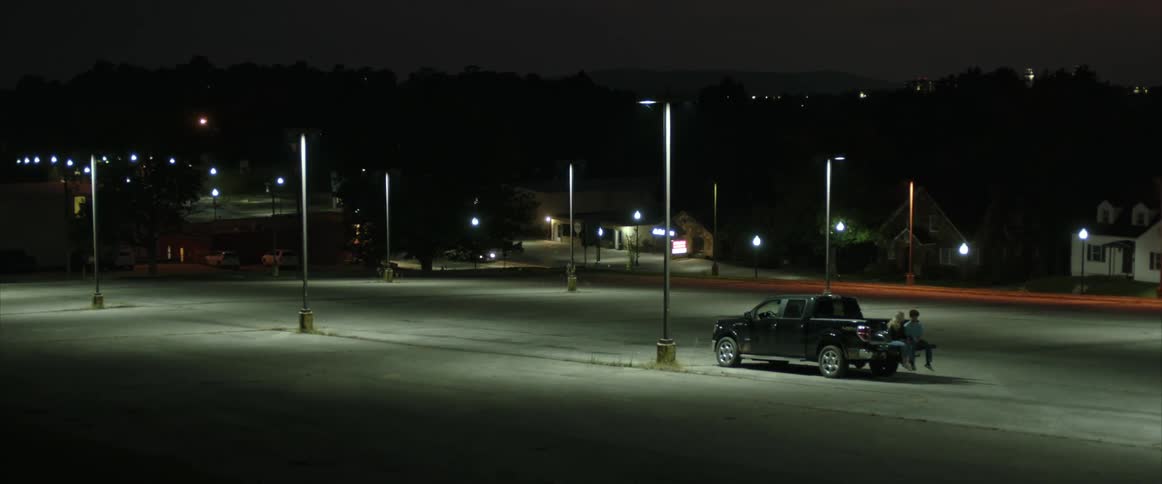 A man and woman talk while sitting together in the bed of a truck in an empty parking lot at night