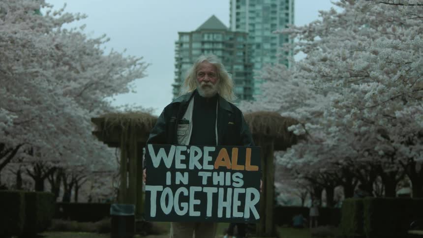 An elderly man holding a protestor sign looks into the camera while standing in a public park in a city during the day