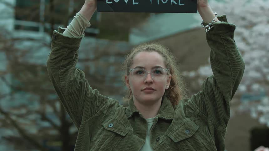 Blonde female protestor with ponytail glasses and green jacket holds up sign