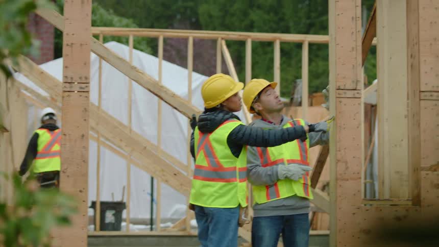 A construction worker is measuring a beam at a construction site when a female walks over and speaks to him and pats him on the back