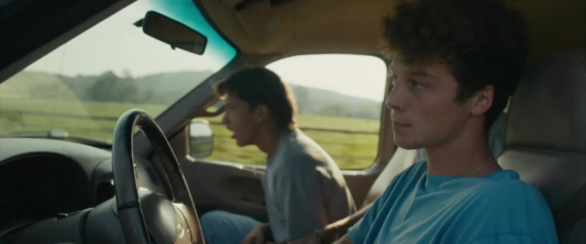 A teenage boy is driving a car with two other teenagers and they play around as the car speeds down a road