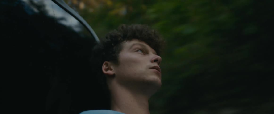 A young man looks up at the sky while sitting in the bed of a truck as it travels through a woodland during the day