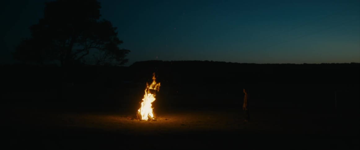 A person is standing in the darkness watching a bonfire burn