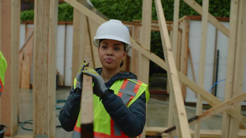 A female construction worker is carrying a piece of wood through a construction site and she waves to a coworker