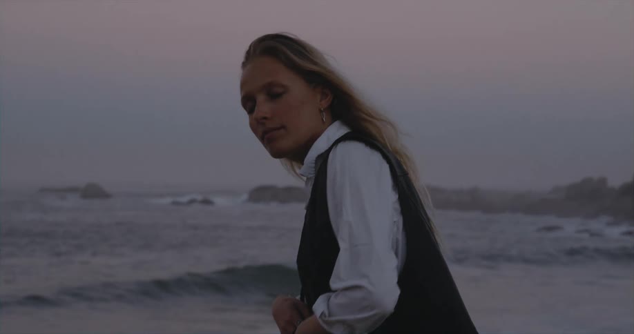 Blonde woman with white shirt and black waistcoat dances on the beach at dusk
