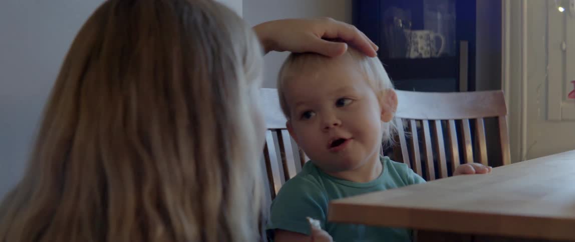 A woman affectionately rubs her young son's head while he eats at a table.