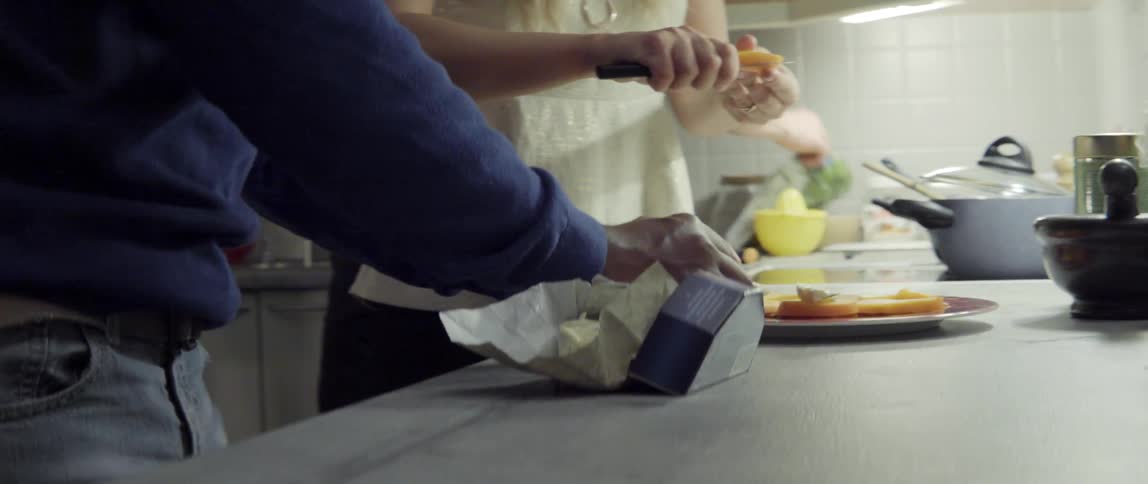 A woman and man slice cheese and place it on a plate while standing in a kitchen