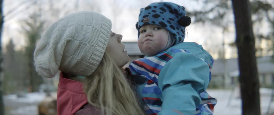 A blonde woman wearing a knit hat kisses her young toddler on a cheek.
