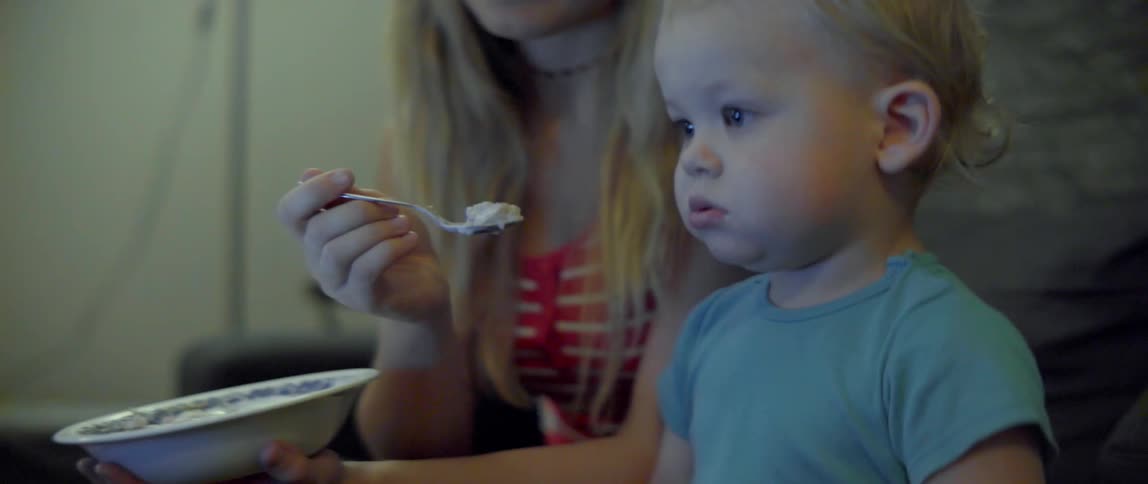 A woman holds a spoon of healthy food for her distracted young son indoors.