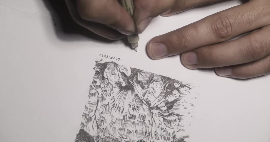 A man is sitting down at a table drawing with a black pen and signing and dating his picture