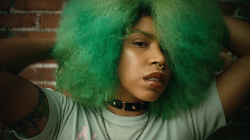 A woman runs her hands through her green hair while posing in front of a brick wall