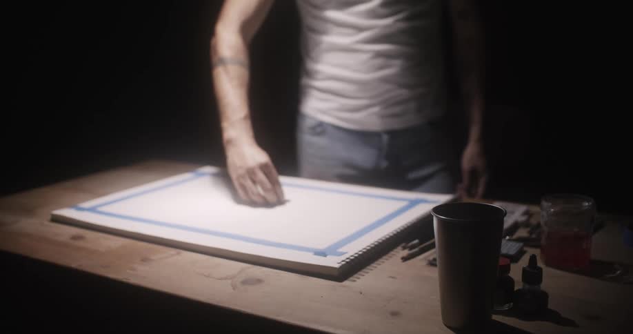 A man is standing at a wooden table and sketching on a canvas