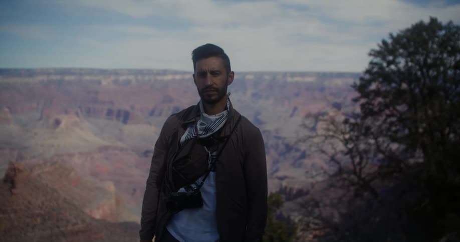 A man looks into the camera while standing atop a vast canyon during the day