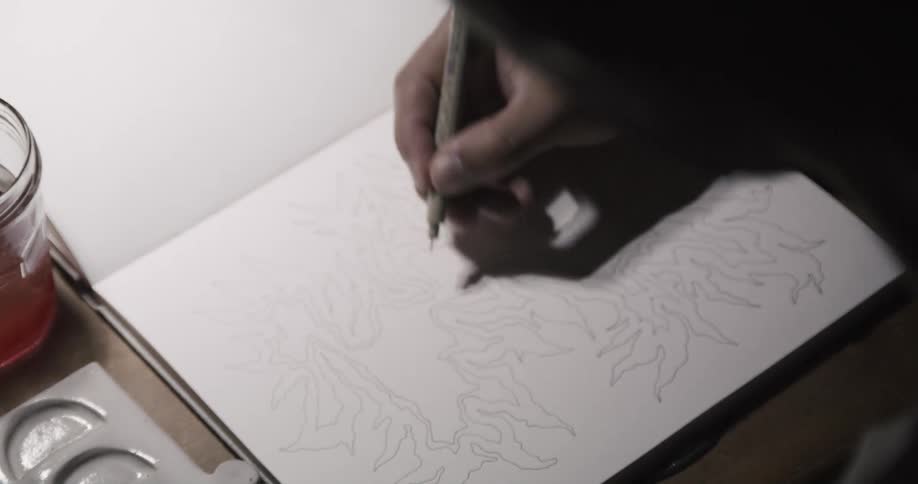 A man sitting at a desk draws in a sketchbook using a pen