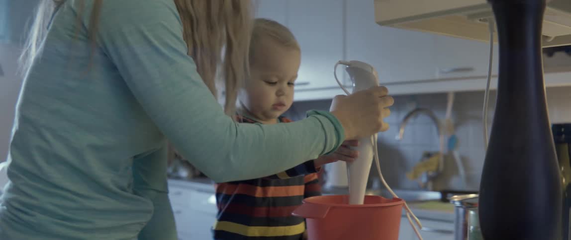 A young toddler dumps ingredients into a red bowl while his mother helps him.