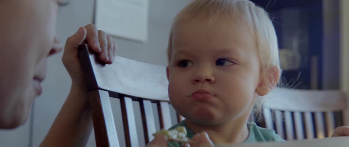 A young toddler smiles happily at his mother while eating a healthy snack indoors.