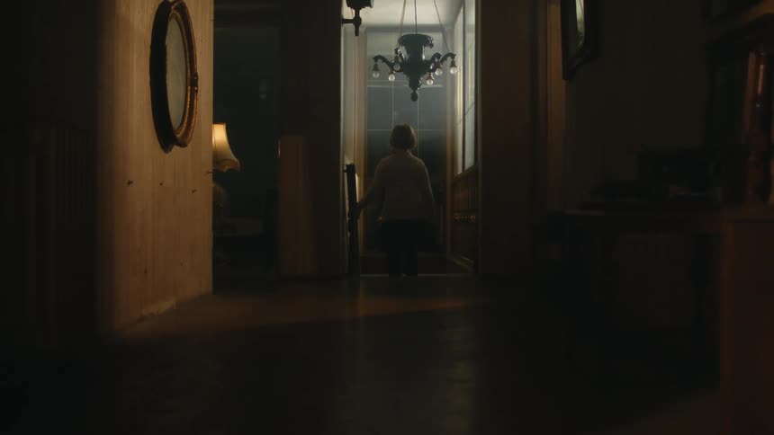 A woman walks through a corridor and descends a staircase in a house at night