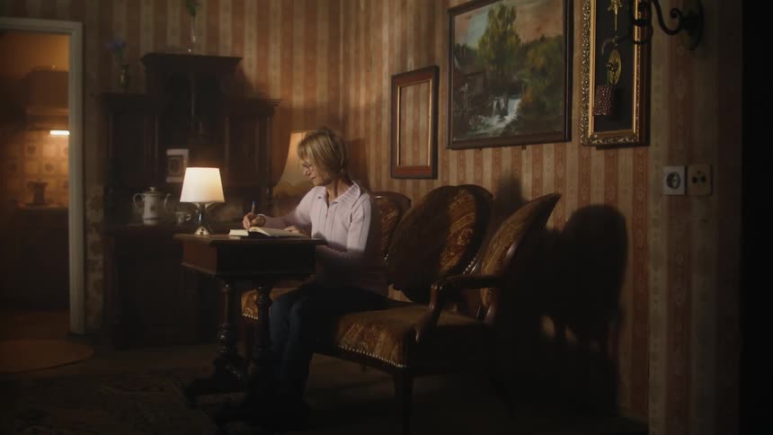 An elderly woman is sitting at a table writing in a book and there is an empty picture frame on the wall behind her