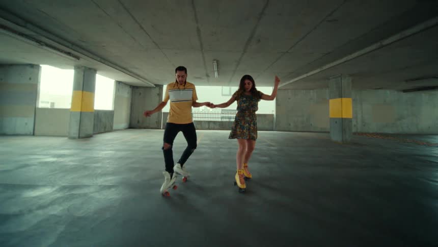 A man and woman hold hands while dancing in roller skates in a parking garage during the day