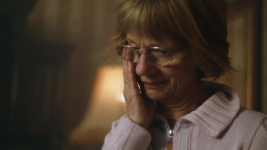 Old woman standing inside her house looking at something with a sad face while starting to cry