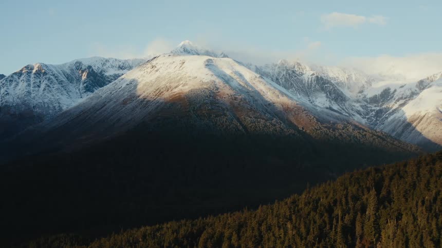 A snow capped mountain ridge looms over smaller mountains covered in forest during the day