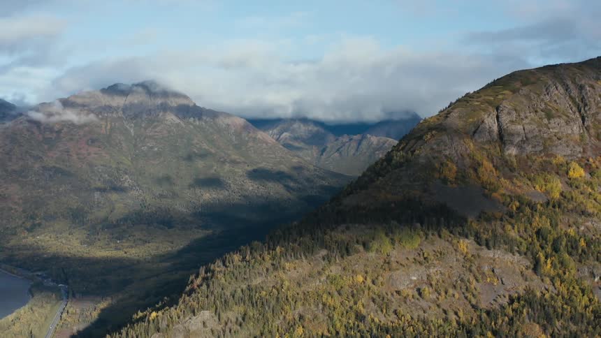 A mountain ridge covered in forest looms over a road running alongside a lake