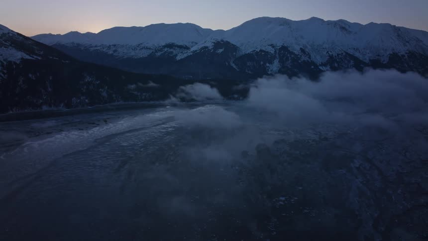Water flows past frozen ground and snow capped mountains as sun sets